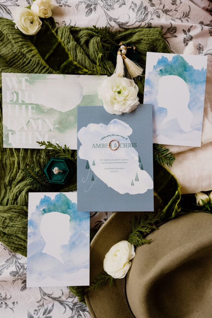 Flat lay of the Invitation suite with the groom’s hat, styling blooms, ring box, wedding band, and earrings with a forest green and emerald color theme. Vermont winter elopement