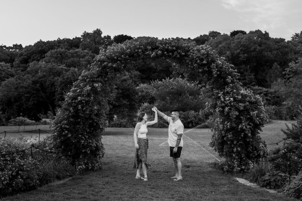 Engaged couple session at the Arnold Arboretum. Diverse Couple, engaged mixed couple, Candid moments of the couple running in the rose garden while the sprinklers are going off in black and white