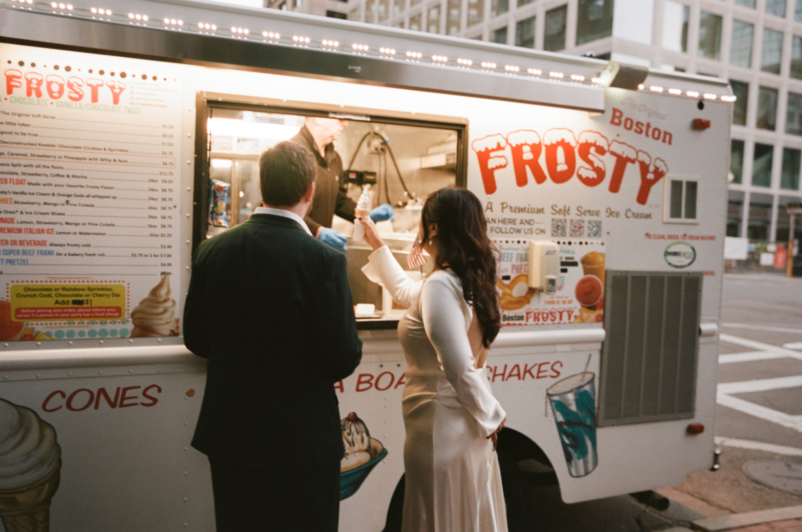 A romantic and candid 35mm film photograph capturing a newlywed couple at a classic Boston Frosty ice cream truck. The bride, in an elegant gown, and the groom, in a suit, share a lighthearted moment as they order treats together. The soft, nostalgic tones of film enhance the charm and intimacy of the scene, perfect for couples seeking a timeless and artistic wedding photographer to document their unique love story.