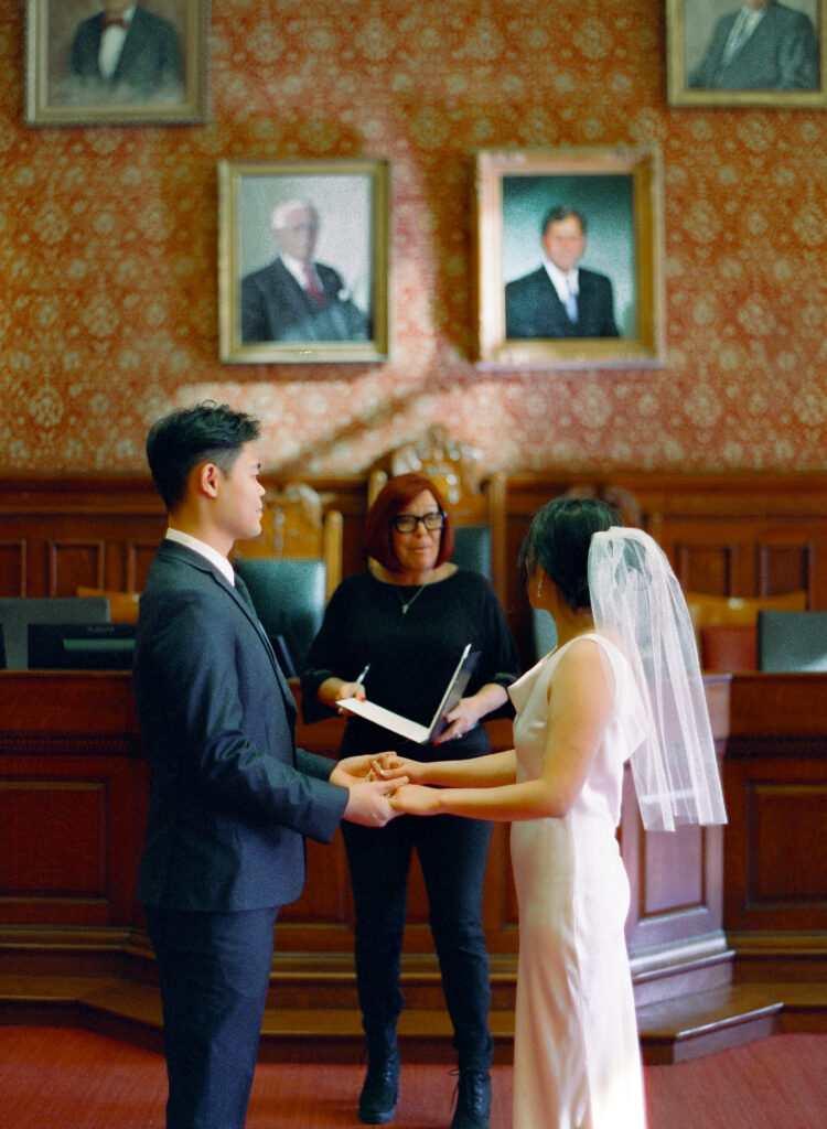 Anna and Huimin standing hand in hand inside Cambridge City Hall as they say “I do” on Valentine’s Day.