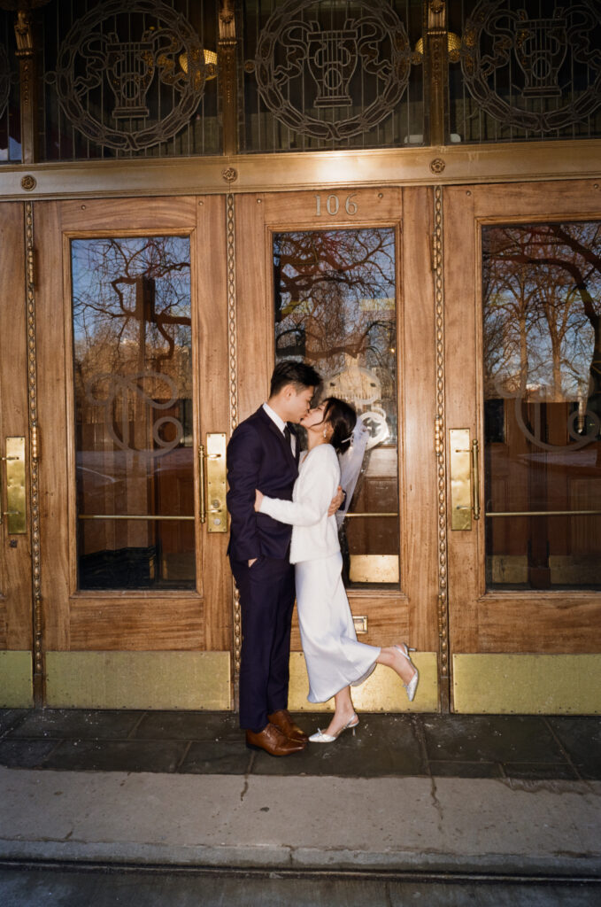 Newlywed couple standing in front of dramatic vintage doors in Boston’s Theater District during their Cambridge City Hall elopement, captured on film by a Boston elopement photographer.