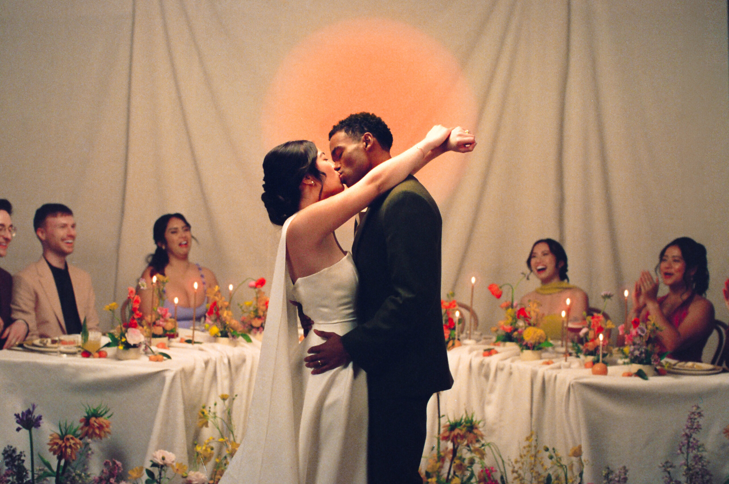 Bride and groom kiss in front of their wedding party during an indoor floral reception, captured on 35mm film by wedding film photographer Castillo Holliday Photo + Film