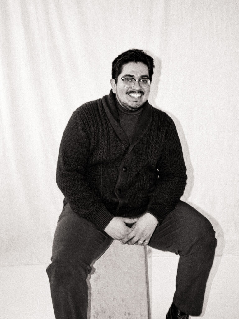 Black and white film photo of Javi from @castillohollidayphotoandfilm smiling while seated on a wooden cube during the Spring 2025 editorial.