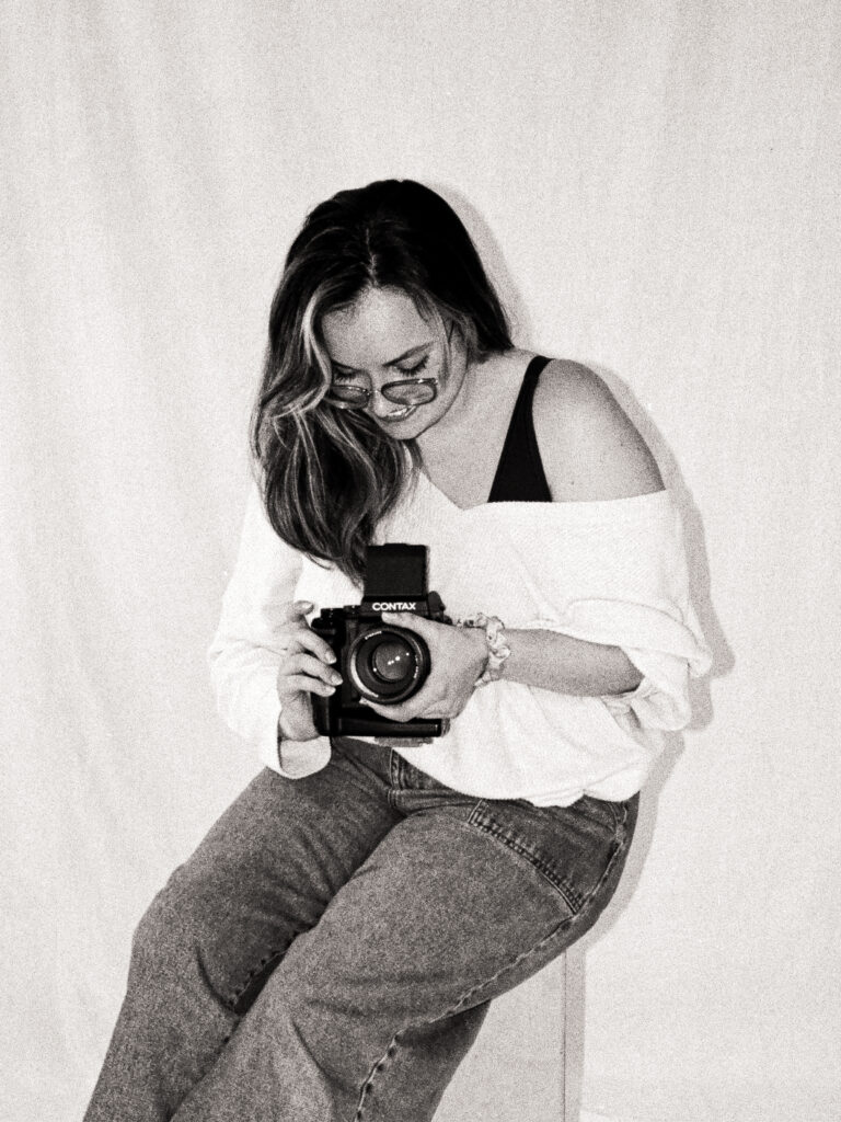 Black and white film portrait of Dominique from @castillohollidayphotoandfilm seated on a stool, smiling down at a Contax 645 medium format camera during the Spring 2025 editorial.