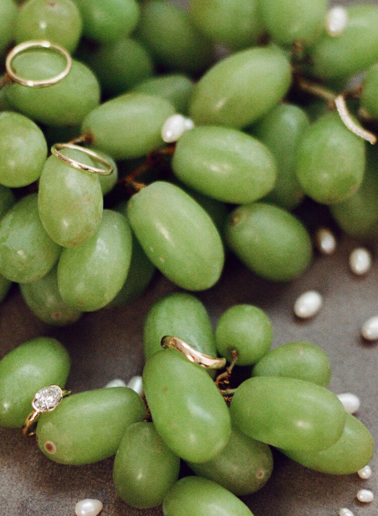A playful flatlay of green grapes adorned with gold wedding bands and a diamond engagement ring. Scattered freshwater pearls add texture and whimsy. A creative, food-inspired take on wedding details from a Spring 2025 film editorial.