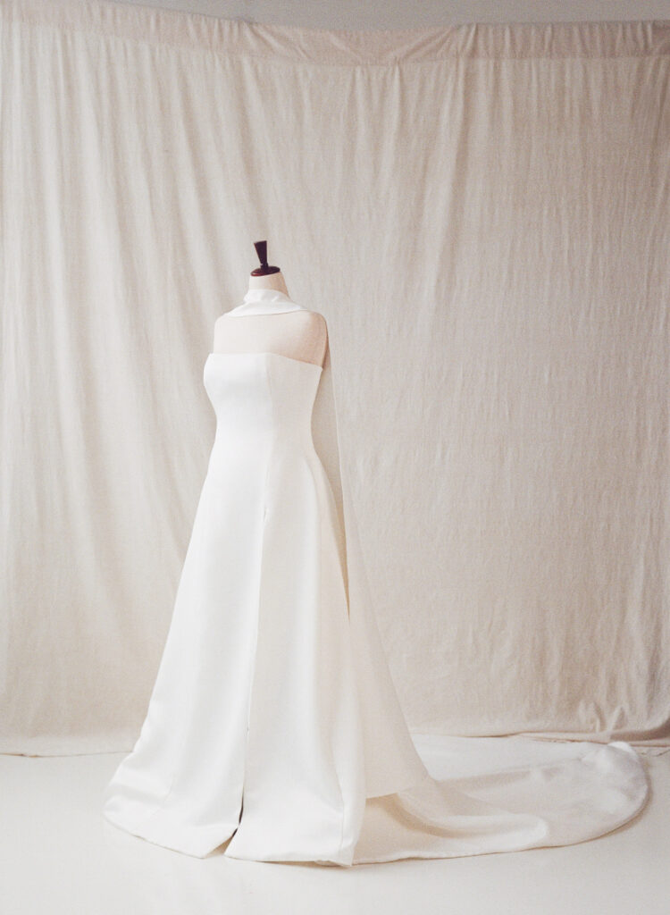 A sleek, strapless wedding gown displayed on a dress form against a soft ivory backdrop. The structured bodice flows into a full A-line skirt with a subtle front slit and long train. A modern, sculptural shoulder wrap adds architectural interest. Photographed on film for a Spring 2025 queer wedding editorial.