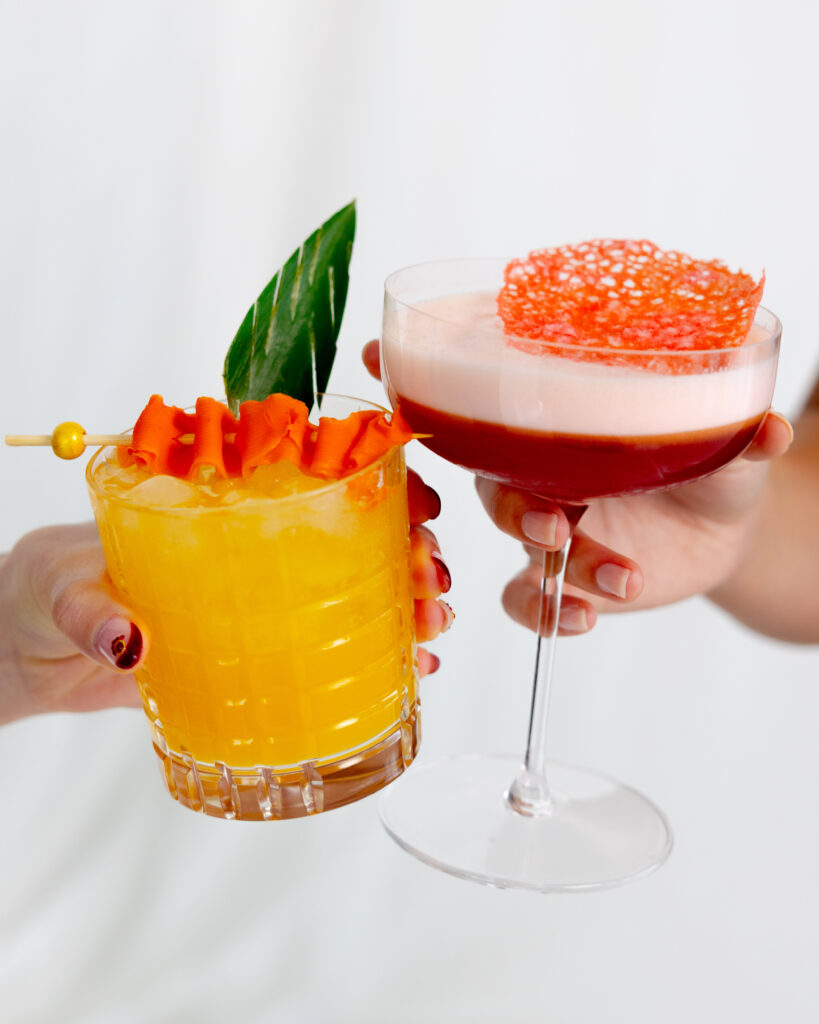 Close-up of two hands holding vibrant signature cocktails—one yellow drink with a leaf and orange garnish, the other a red and pink cocktail topped with an orange tuile. A joyful toast during the Spring 2025 queer wedding editorial, styled with color-forward, celebratory details.