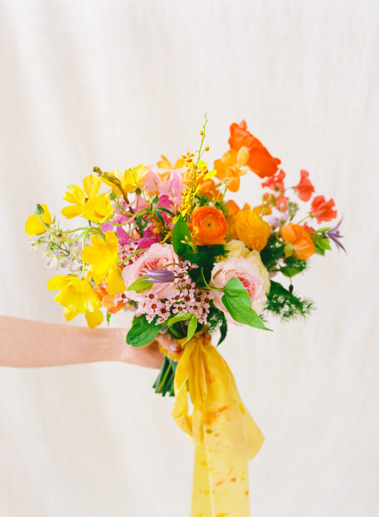 A vibrant spring wedding bouquet held in one hand, featuring butterfly ranunculus, orange ranunculus, pink garden roses, wax flower, and clematis, tied with a hand-dyed yellow ribbon. Captured on medium format film against a soft ivory backdrop.