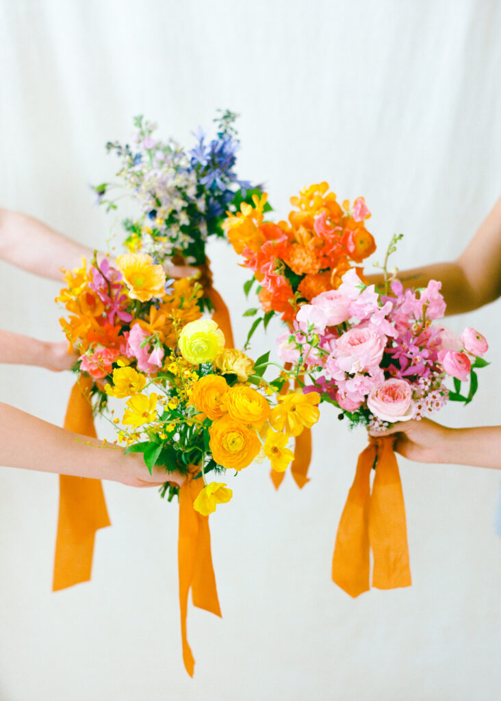 Five colorful spring wedding bouquets held up by different hands, each featuring vibrant florals like ranunculus, roses, delphinium, and orchids, all tied with flowing golden-orange ribbons against a soft ivory backdrop. Captured on medium format film.