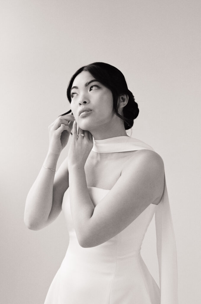Black and white bridal portrait of an Asian bride putting on earrings before her wedding ceremony, wearing a modern satin gown with a structured neckline. Captured on film in a Boston wedding editorial focused on fashion-forward styling and diverse representation.