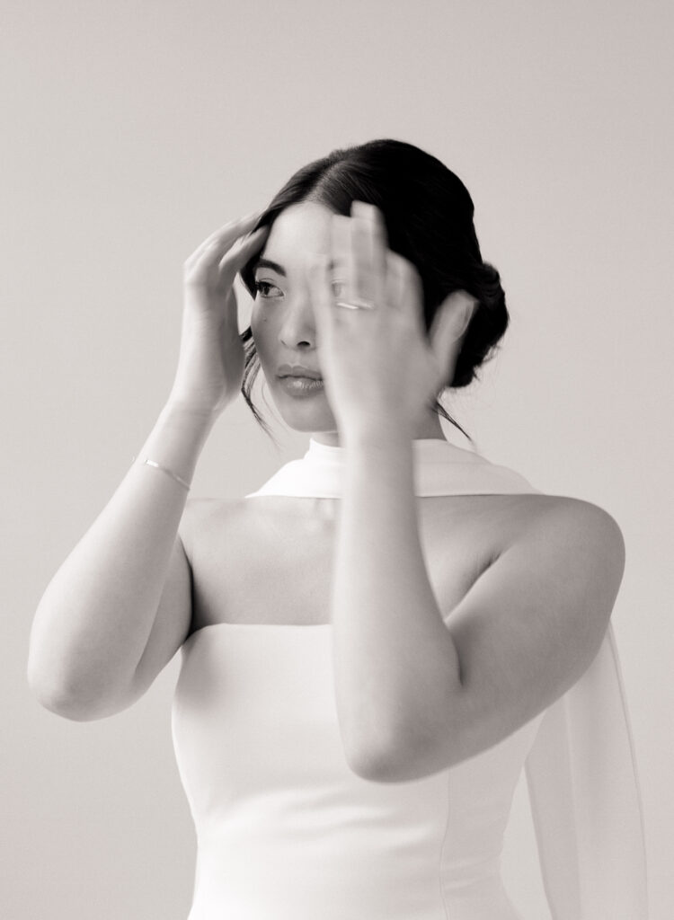 Black and white bridal portrait of an Asian woman adjusting her hair before her wedding ceremony, wearing a minimalist satin gown with a modern cape neckline. A timeless and elegant Boston wedding editorial captured on film, perfect for couples seeking high-end film wedding photography.