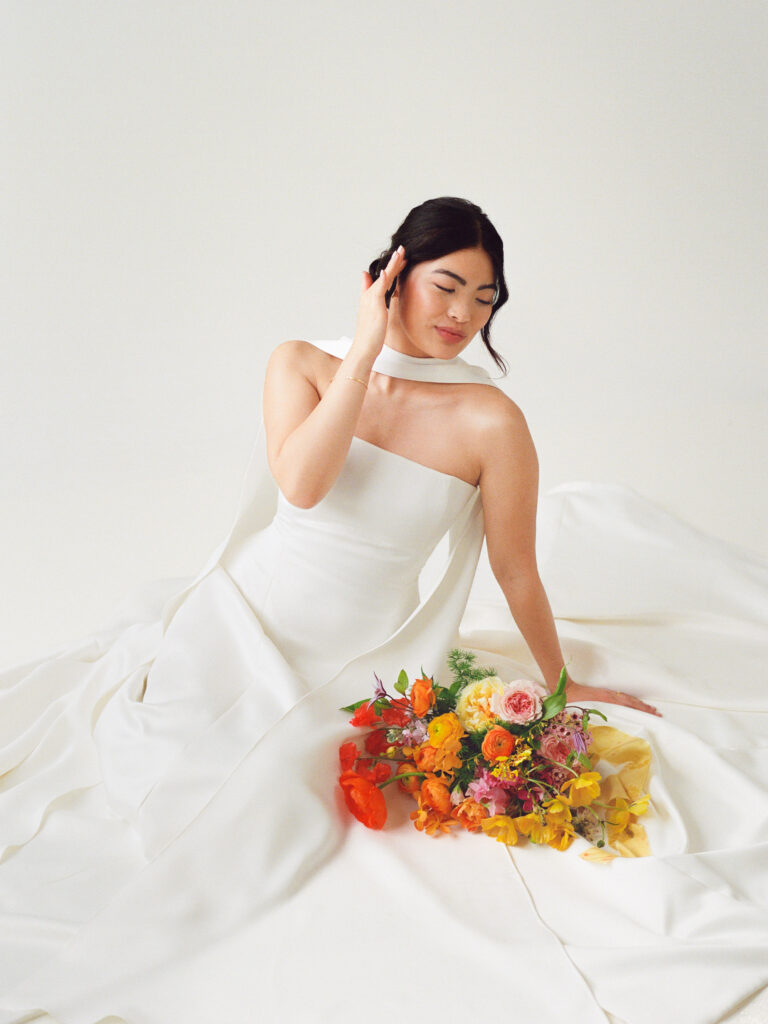Modern Asian bride in a minimalist white wedding gown seated on the floor with a vibrant spring bridal bouquet. Editorial wedding portrait captured on film in a Boston studio featuring colorful floral design by Rumphius Farms.