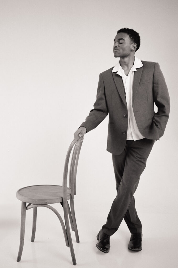 Full-body black and white film portrait of a groom in a relaxed pose, leaning on a wooden chair in a classic suit and open-collar shirt. Editorial groom style captured on 35mm by a Boston-based film wedding photographer for a modern spring wedding inspiration shoot.