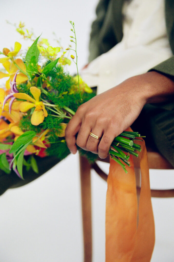 Close-up of a groom’s hand holding a bright spring bouquet with yellow orchids and greenery, wrapped in orange silk ribbon. His gold wedding band adds a subtle touch of elegance. Captured on film by a Boston-based film wedding photographer for a modern editorial shoot.
