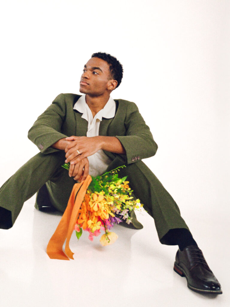 Modern groom in a tailored olive green suit sitting on the floor with a colorful spring wedding bouquet featuring orchids, tulips, and ranunculus. Captured on film by a film wedding photographer in Boston for a vibrant editorial wedding inspiration shoot.