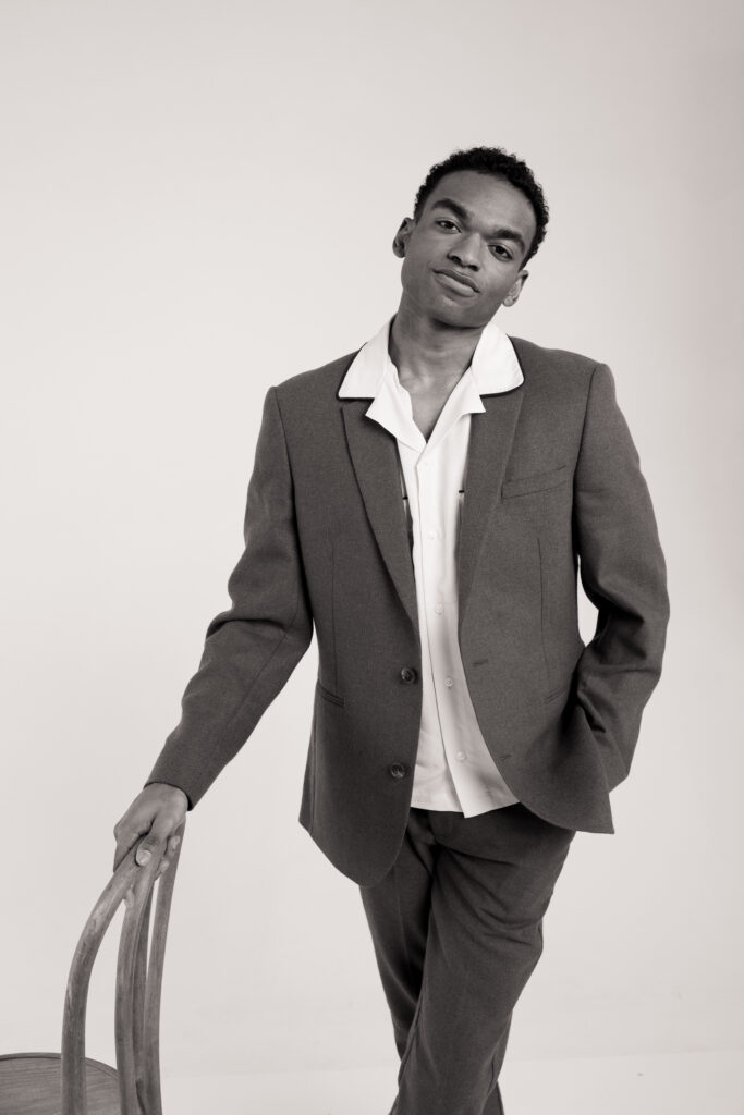 Black and white portrait of a stylish groom in a tailored suit with a relaxed open collar shirt, leaning on a vintage bentwood chair. Modern groom fashion inspiration for diverse spring weddings, captured on 35mm film by a Boston-based film wedding photographer.