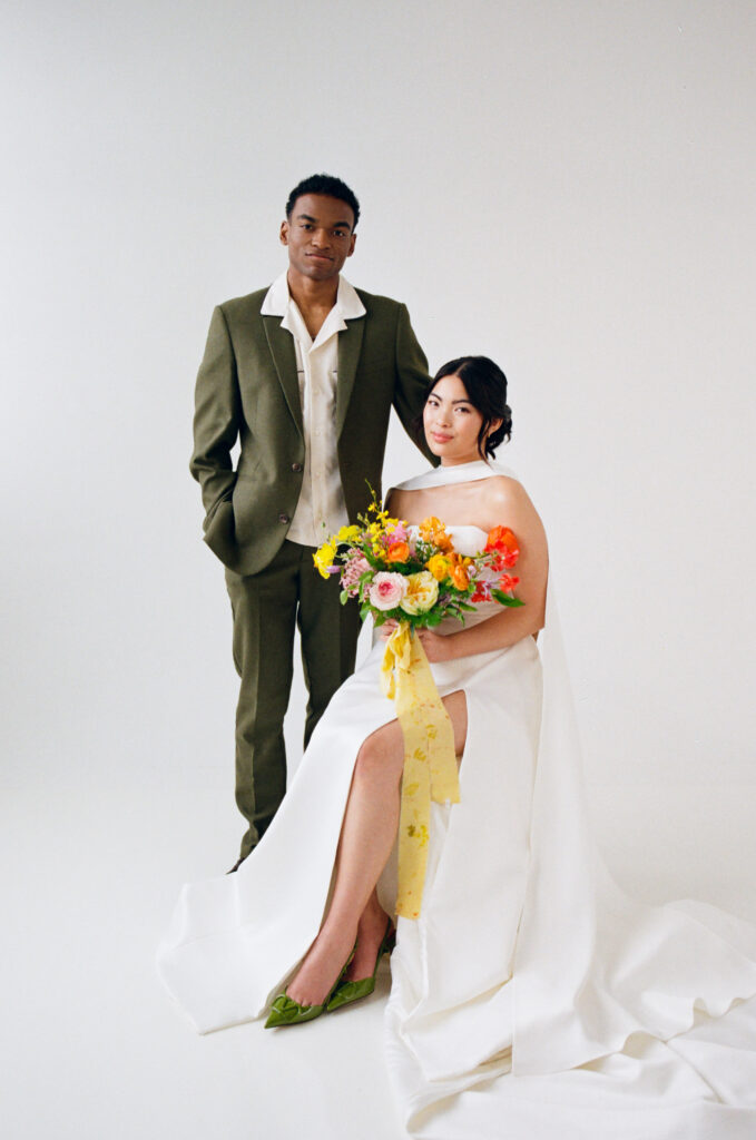 A modern couple poses for a timeless studio wedding portrait. The bride sits in a structured white gown with a high slit and flowing scarf detail, holding a vibrant spring bouquet with yellow ribbon. Her partner stands beside her in an olive green suit with a relaxed collared shirt. Captured on film by a Boston film wedding photographer during a minimalist editorial shoot.