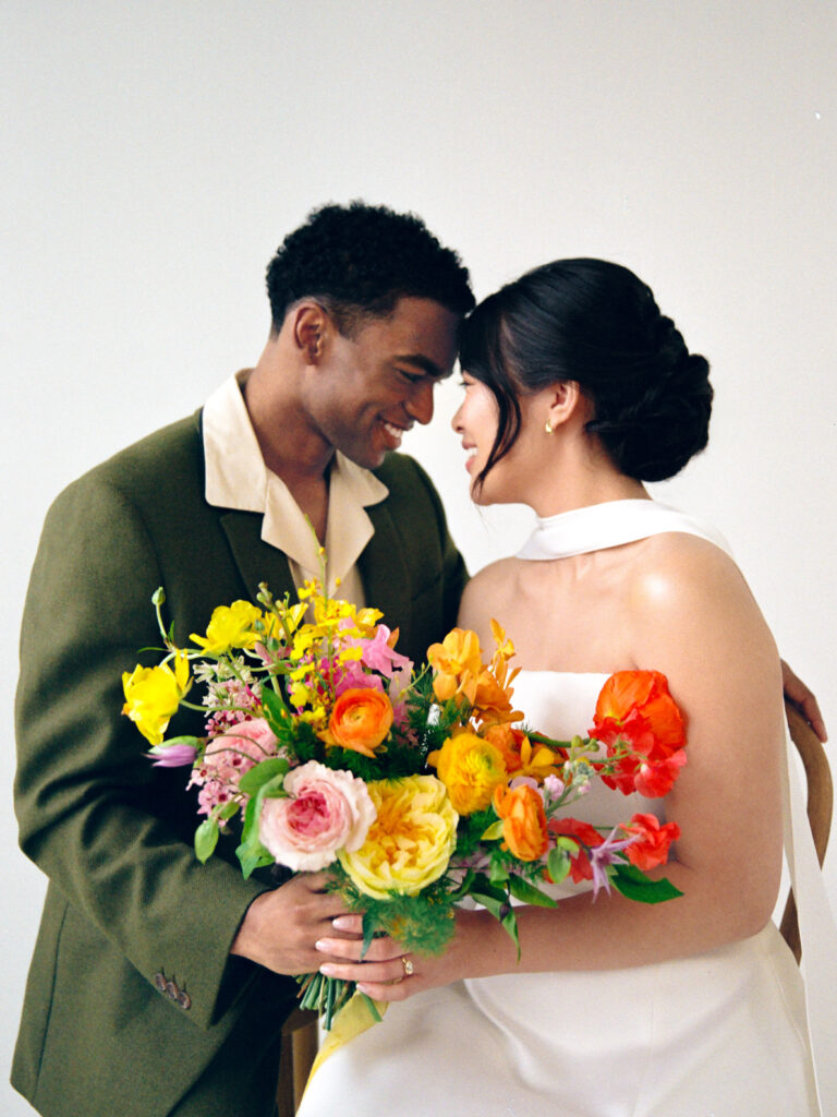A joyful wedding couple shares an intimate forehead-to-forehead moment in a bright studio. The bride, in a strapless white gown, holds a bold spring bouquet bursting with yellow, orange, and pink blooms. Her partner, dressed in a relaxed olive suit, gently wraps his hand over hers. Captured on film by a New England film wedding photographer during a minimalist editorial session.