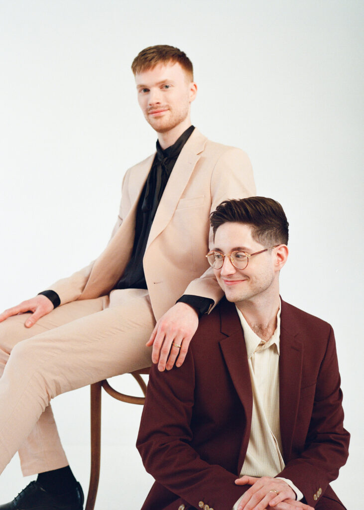 A stylish queer couple pose together as wedding guests, wearing modern suits in blush and burgundy tones. One partner sits on a bentwood chair while the other kneels at his side, both smiling softly. Captured on film by a Boston film wedding photographer during a minimalist editorial studio wedding shoot.