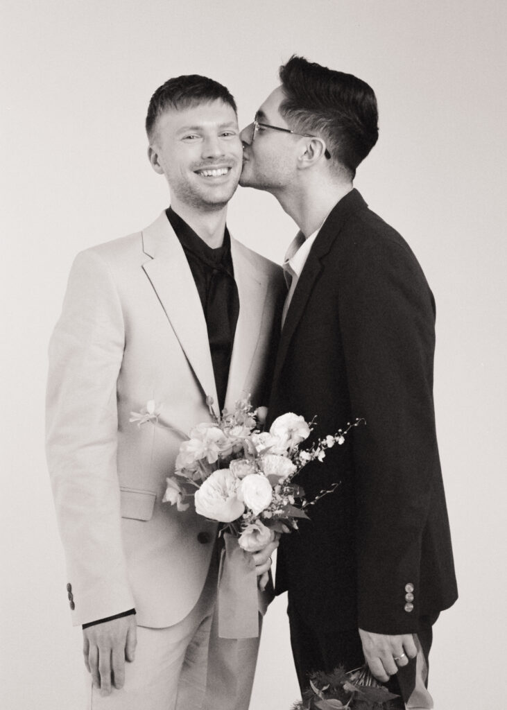 A joyful queer couple, dressed as wedding guests in tailored suits, share an affectionate moment as one kisses the other on the cheek. They each hold spring wedding bouquets. Captured on black and white film by a Boston film wedding photographer during a minimalist studio editorial.