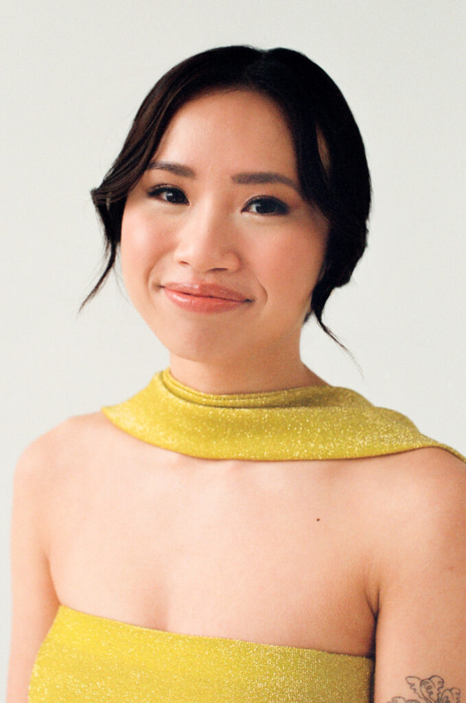 A guest smiles softly while wearing a shimmery golden yellow gown with a structured neckline and shoulder wrap detail. Her hair is styled in a low, elegant updo with delicate face-framing strands, and a subtle floral tattoo peeks out from her arm. Captured on film as part of a Spring 2025 wedding editorial centered on modern, inclusive celebration in Boston.