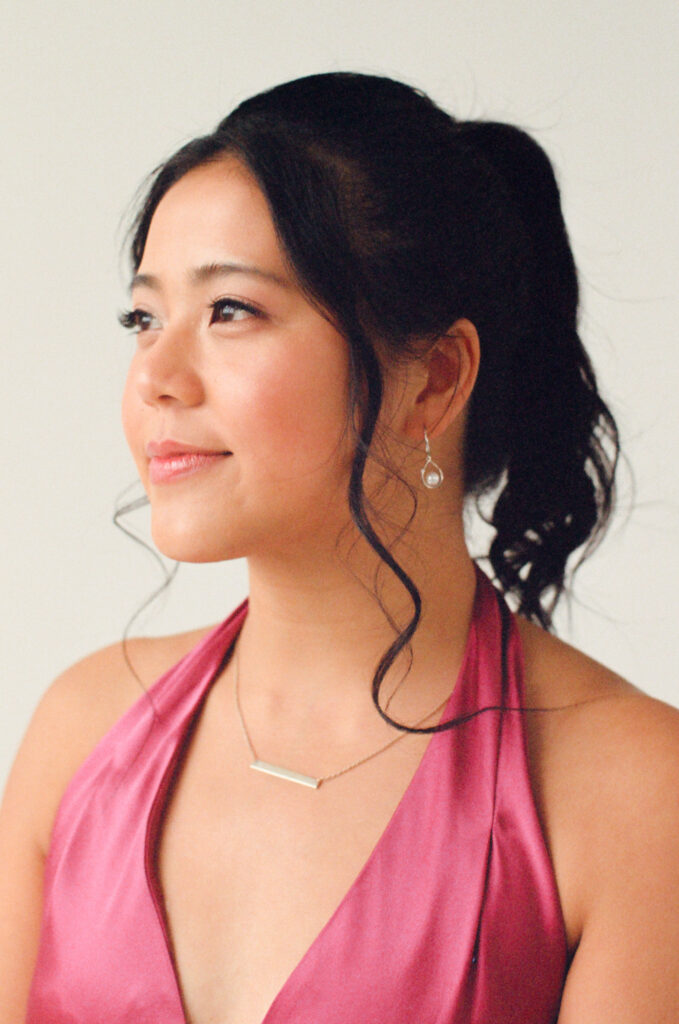 A wedding guest gazes softly to the side with a peaceful smile, wearing a magenta satin halter gown. Her hair is styled in a high ponytail with face-framing curls, paired with drop earrings and a minimalist bar necklace. Captured on film during a Boston-based Spring 2025 editorial celebrating diverse wedding inspiration.