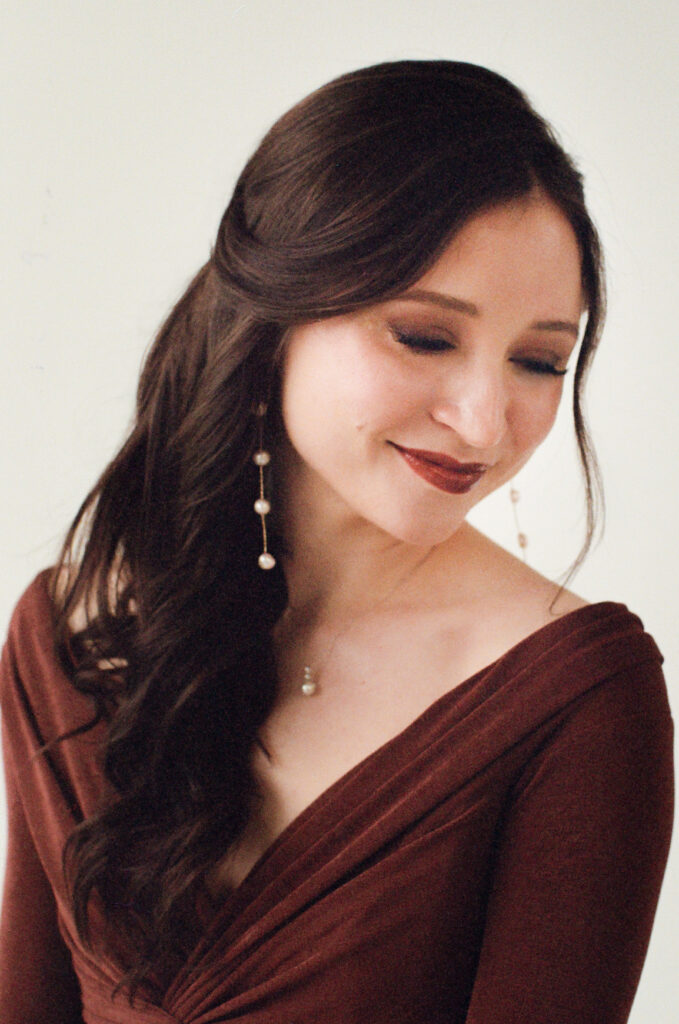 A wedding guest with long, softly curled brunette hair smiles with her eyes closed in a serene moment. She wears a deep burgundy velvet gown with a draped neckline, pearl drop earrings, and a delicate pearl necklace. Captured on film by a Boston film wedding photographer during a Spring 2025 editorial focused on inclusive wedding inspiration.