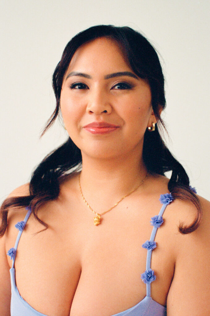 A close-up portrait of a guest from the Spring 2025 editorial, smiling softly with confidence. She wears a lavender dress with floral appliqué straps, gold hoop earrings, and a delicate gold necklace with a heart-shaped charm. Captured on film by a Boston-based film wedding photographer in a sunlit studio setting.