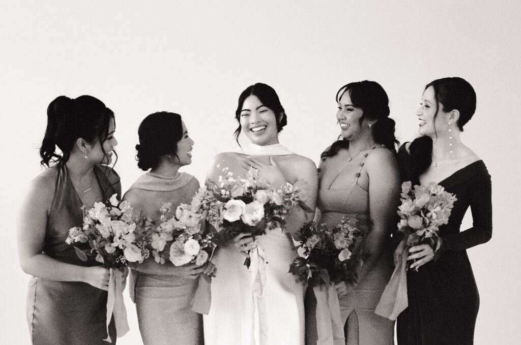 A joyful bride stands at the center of her bridal party, surrounded by five guests holding lush bouquets. Everyone is laughing and looking toward each other in a moment of genuine connection. The group wears a palette of mixed spring tones—berry, gold, mauve, and deep plum—while the bride glows in a structured, modern white gown. Captured on black and white film as part of a Spring 2025 wedding editorial rooted in community, celebration, and style.