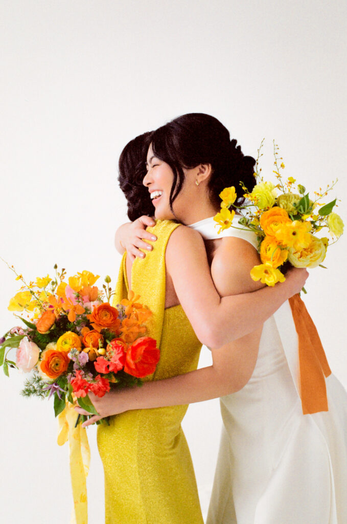 The bride and a guest share a heartfelt hug, both beaming with laughter and joy. The guest wears a vibrant yellow gown and holds a colorful bouquet of orange ranunculus, poppies, and blush blooms. The bride, in a modern white dress with a structured neckline, carries a bouquet of golden yellow florals with an amber ribbon. A radiant moment of connection captured on film during the Spring 2025 wedding editorial.