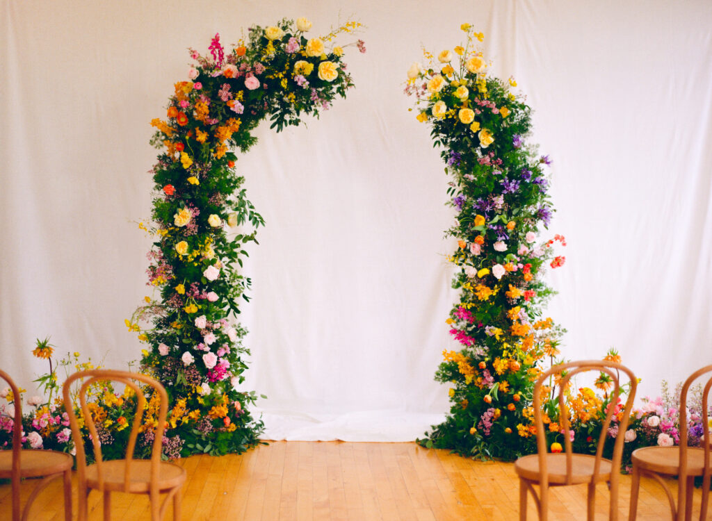 Asymmetrical floral arch installation in a soft studio setting, overflowing with butterfly ranunculus, roses, sweet peas, and wildflowers in vibrant yellow, peach, lavender, and pink tones. Wooden bistro chairs are arranged for an intimate ceremony on honey-colored floors. Captured on medium format film.