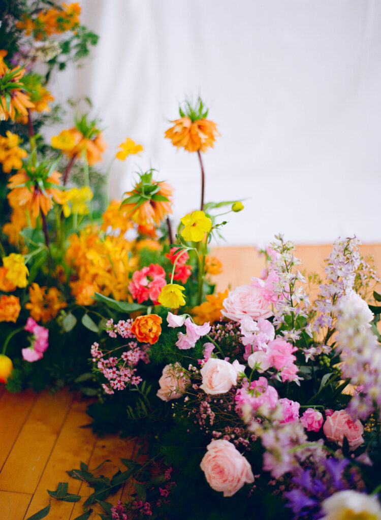 Lush spring floral installation featuring a mix of pastel pink roses, lilac delphinium, yellow poppies, and crown imperials, arranged on hardwood floors with a soft white fabric backdrop, captured on medium format film.