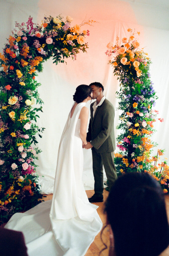 Studio wedding inspiration featuring a couple kissing beneath a bold floral arch by Rumphius Farms. Captured on 35mm film by Boston film wedding photographer Castillo Holliday Photo + Film for a diverse spring wedding editorial.