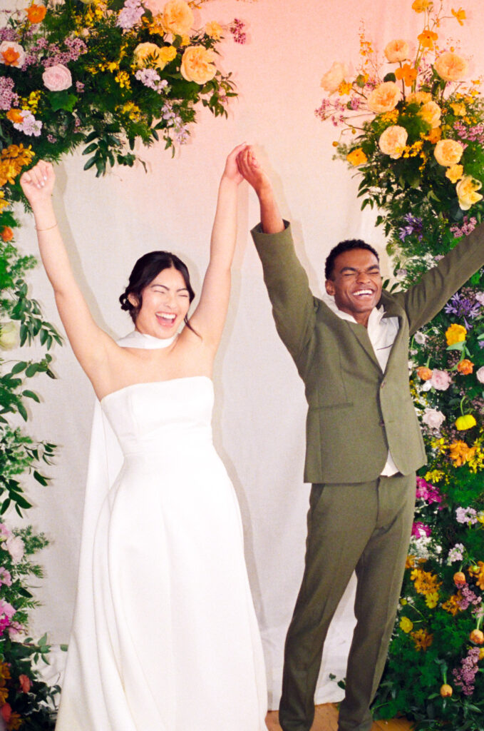 Joyful wedding editorial moment with a stylish couple celebrating beneath a colorful spring floral arch by Rumphius Farms. Captured on 35mm film in a Boston studio by film wedding photographer Castillo Holliday Photo + Film.