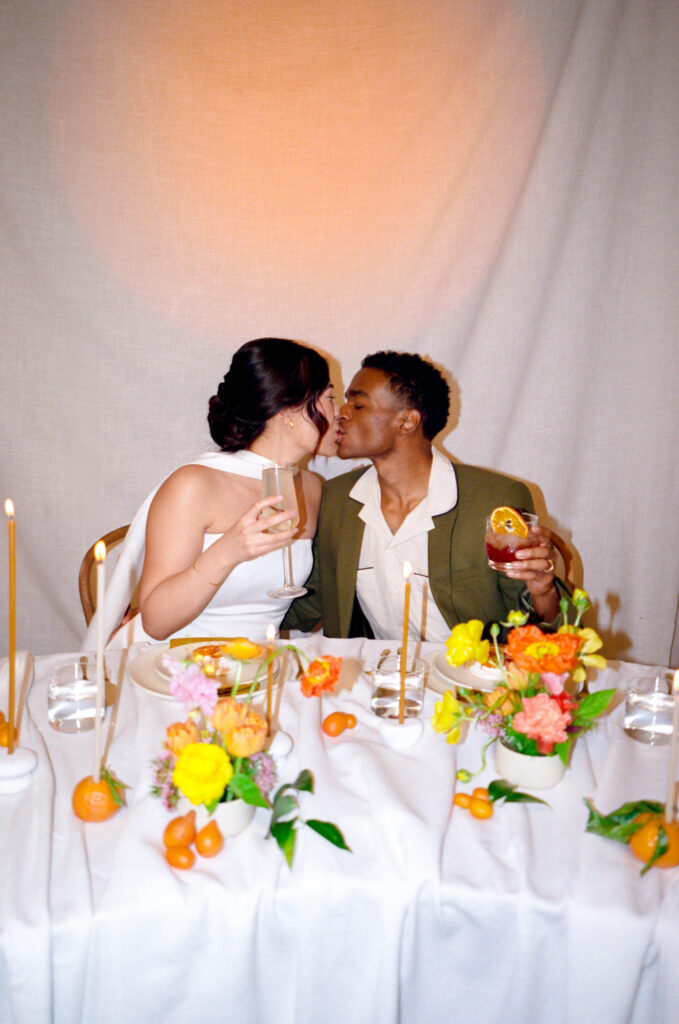 Modern spring wedding inspiration captured entirely on film. A joyful couple shares a kiss at their sweetheart table, surrounded by vibrant citrus florals, taper candles, and handcrafted cocktails. Boston-based film wedding photographer