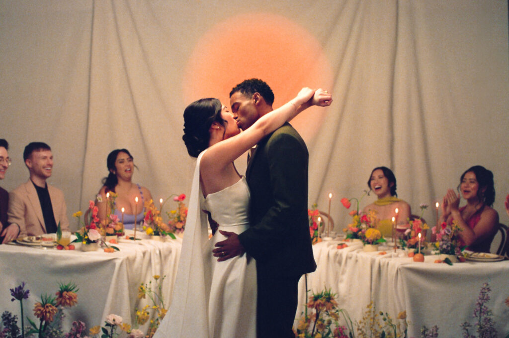 Modern wedding reception inspiration in a Boston studio featuring a joyful newlywed couple kissing in front of their wedding party. Surrounded by vibrant spring florals, colorful taper candles, and a painterly sunset backdrop. Captured on film by New England film wedding photographer Castillo Holliday Photo + Film.