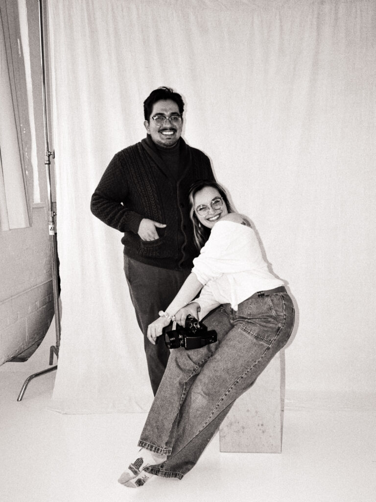 Black and white film portrait of Dominique and Javi of Castillo Holliday Photo + Film. Dominique sits on a wooden stool holding a 35mm camera, smiling warmly in a casual sweater and jeans. Javi stands beside her, wearing glasses and a dark cardigan, both laughing in a sunlit studio with a soft backdrop behind them.