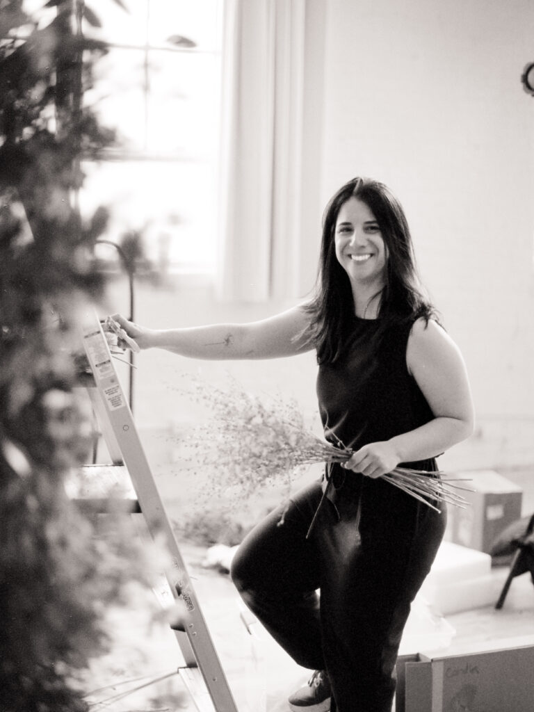 Roxy from @rumphiusfarms smiling while standing on a ladder during floral installation, holding a bundle of flowers in a naturally lit studio space