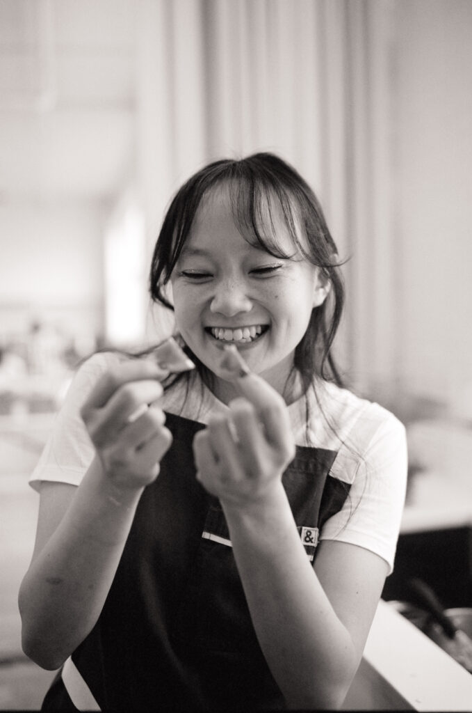 Black and white film portrait of Gen from @genhuang smiling joyfully while preparing a dessert sample during the Spring 2025 editorial.