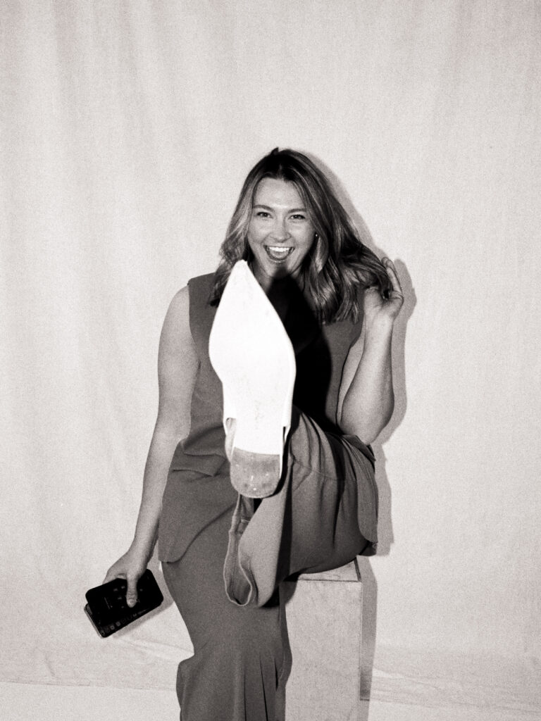 Black and white film portrait of Caroline from @carolinecarletonsocials, captured mid-laugh while playfully kicking up her boot and holding her phone during the Spring 2025 editorial.
