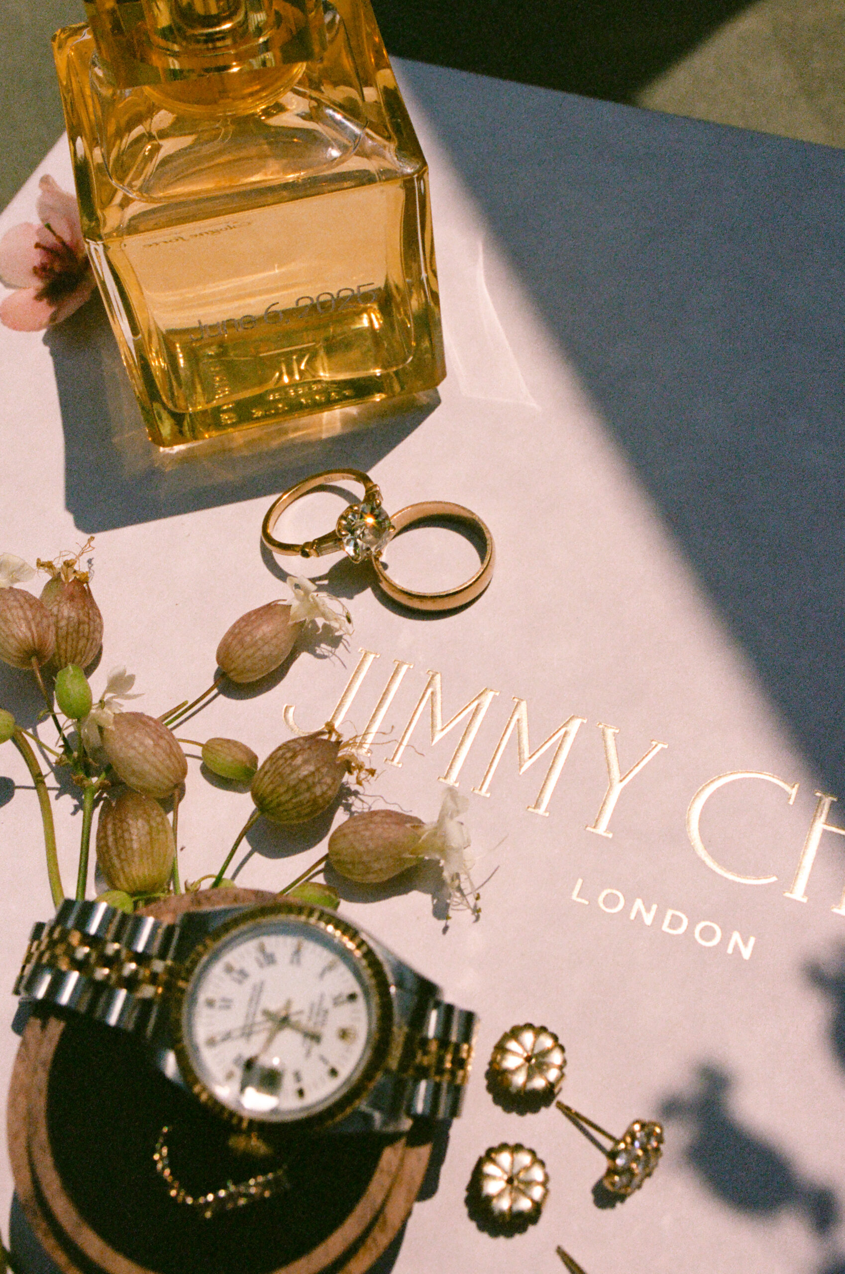 Gold wedding rings, Rolex watch, and Dolce & Gabbana perfume styled with delicate flowers — elegant bridal flatlay detail.