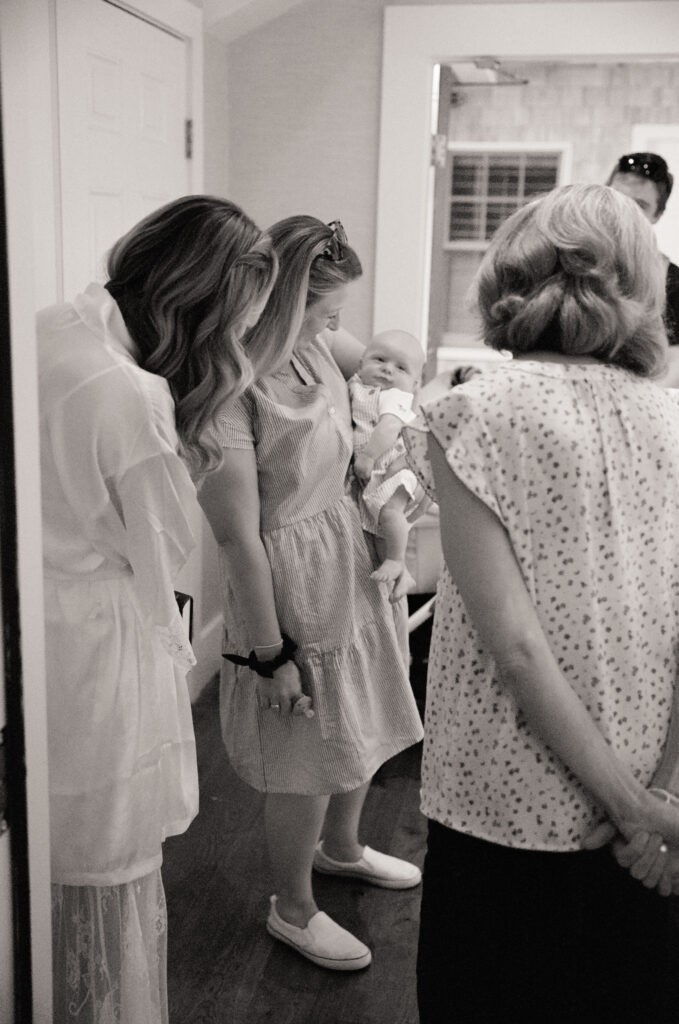 Family members gather around holding a baby during wedding morning preparations – candid black and white film wedding photo.