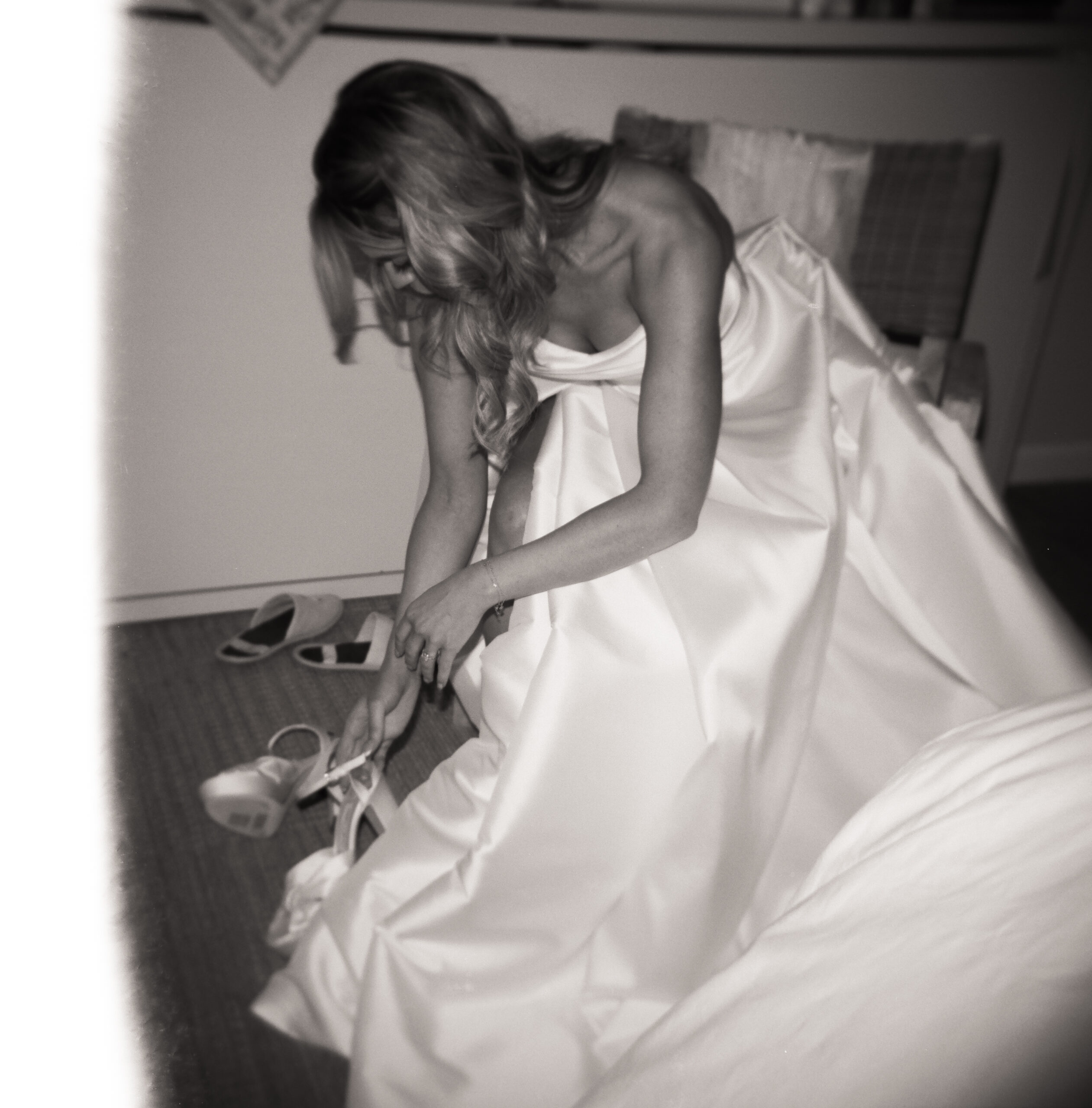 Candid black and white film wedding photo of a bride adjusting her shoes while wearing a flowing white gown, captured in an intimate getting-ready moment.