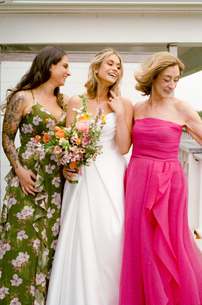 Candid porch moment at a Martha’s Vineyard wedding with the bride Olivia, her mother in a magenta dress, and her sister Dana in an olive green floral dress. The three share a laugh as the mother sets down her glasses, arms loosely around each other, with Olivia holding her bouquet in the breeze.