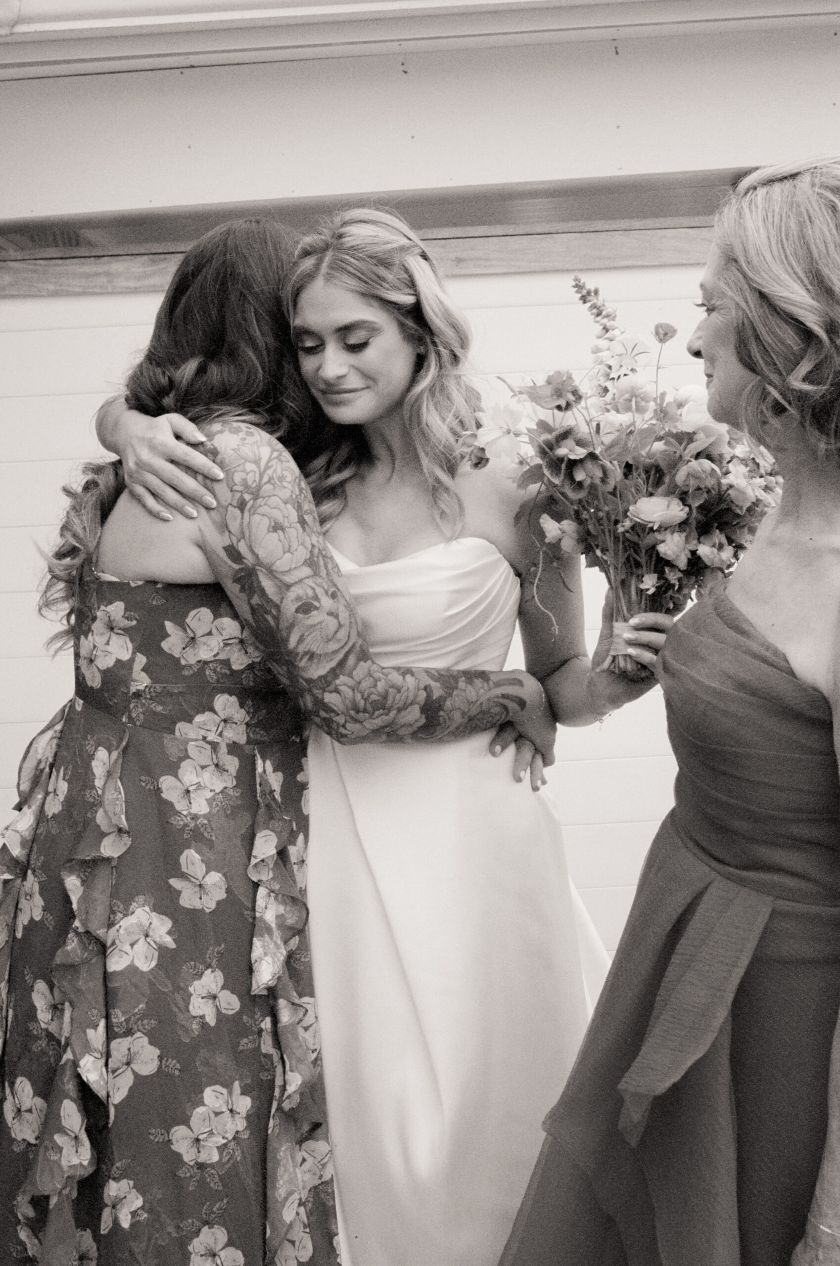Bride embraces a bridesmaid in a floral gown while holding her bouquet, surrounded by loved ones during intimate wedding morning moments.