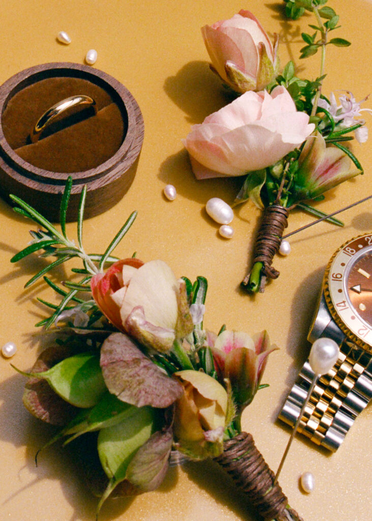 Detail shot of blush pink boutonnières, gold wedding band in a wooden ring box, and wristwatch styled on a mustard background.