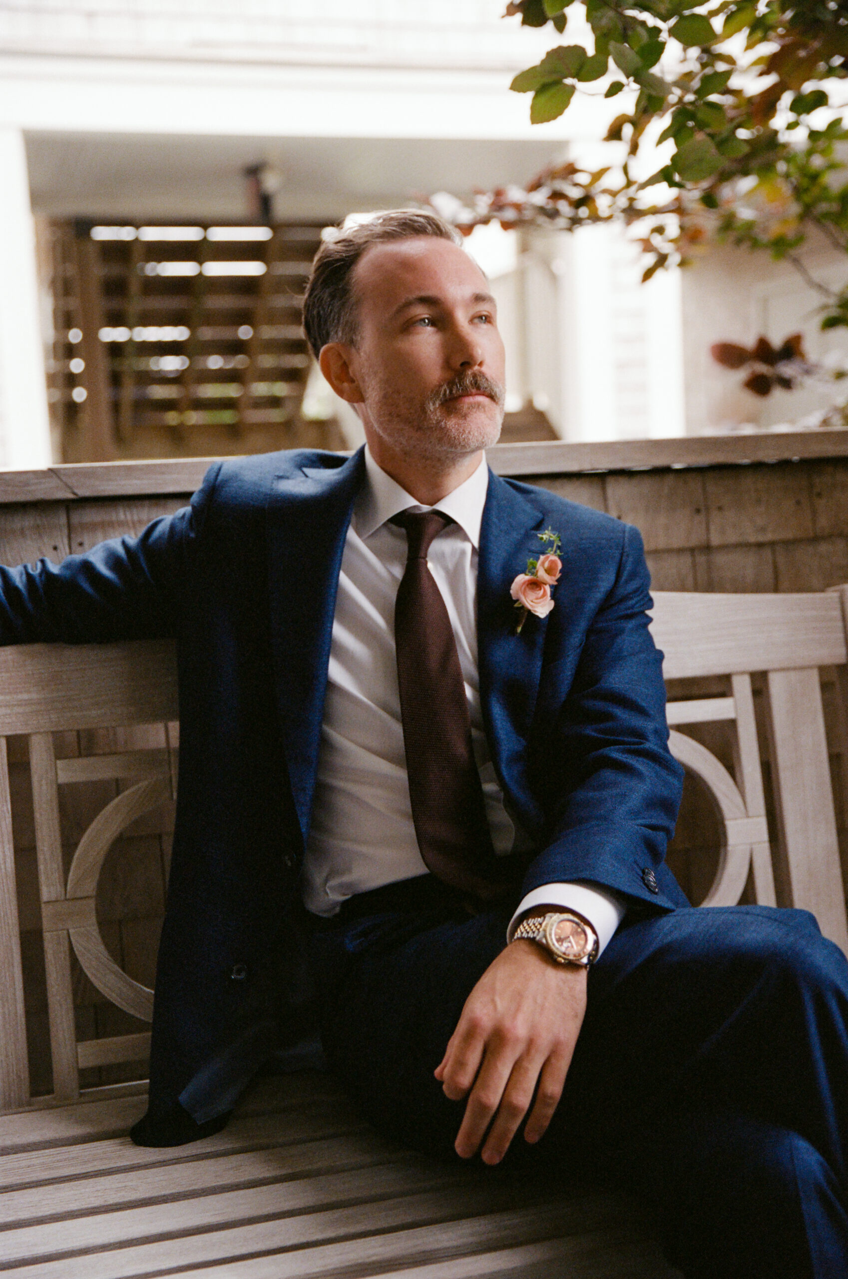Groom in a navy suit with boutonnière poses on a wooden bench, captured on 35mm film before his Martha’s Vineyard wedding.