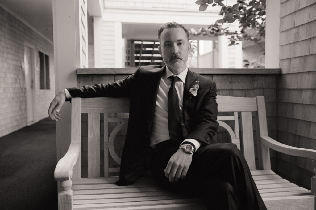 Black and white portrait of the groom sitting confidently on a wooden bench before his Martha’s Vineyard wedding.