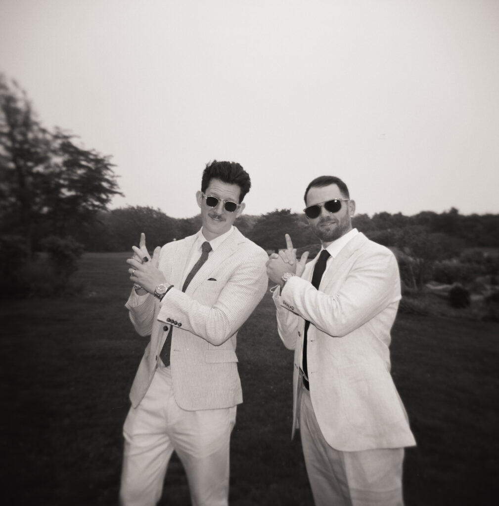 Black and white Holga 120 film photo of two wedding guests in cream suits and sunglasses striking playful finger-gun poses before the ceremony at a Martha’s Vineyard wedding, cinematic and stylish with fog and trees in the background.