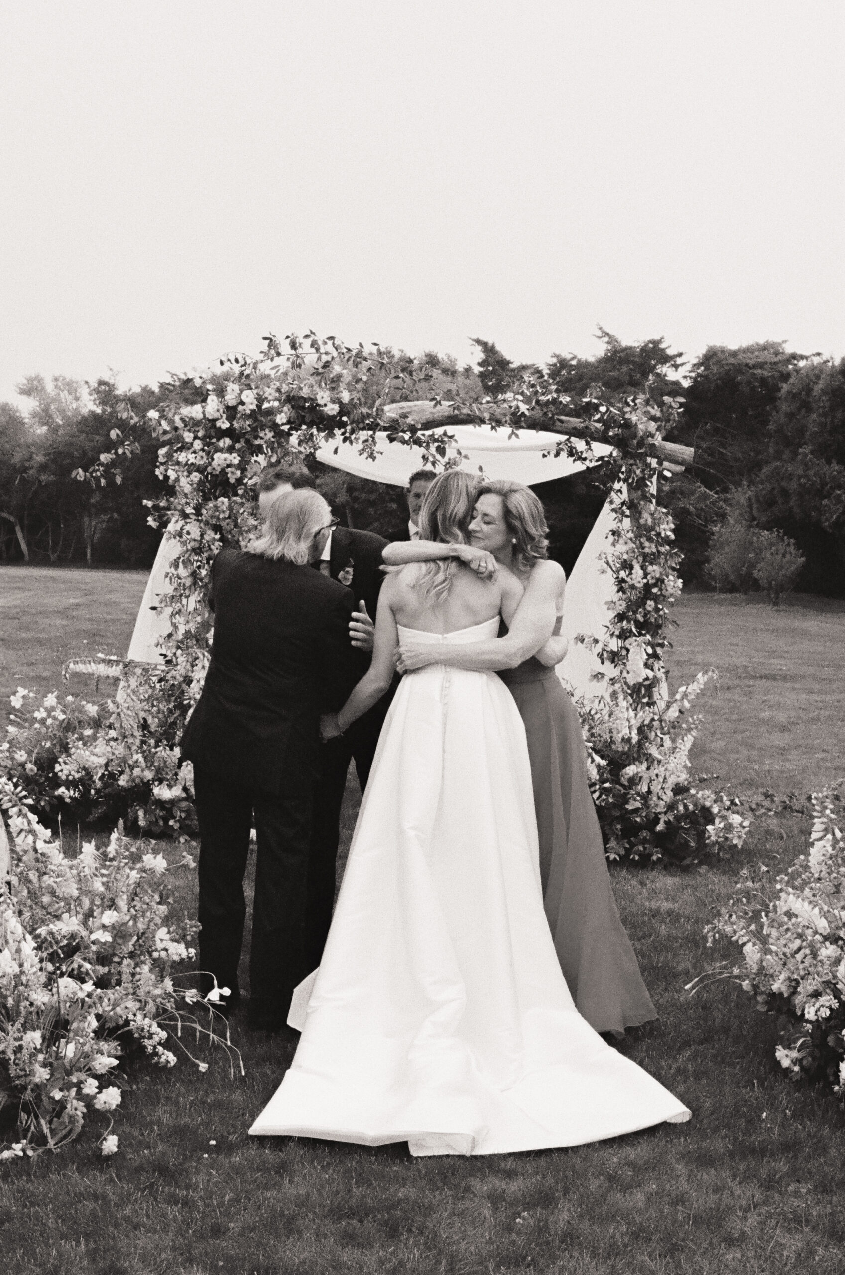 Bride hugging her parents under floral arch during ceremony – candid black and white wedding film photo.